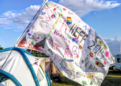 Photo of a flag flying from a tent. The flag is hand decorated with felt tips and depicts HEFF2019 festival