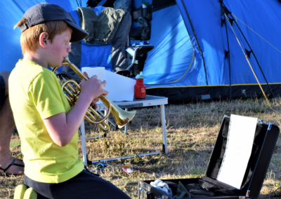 A young child kneeling outside a tent and playing the trumpet