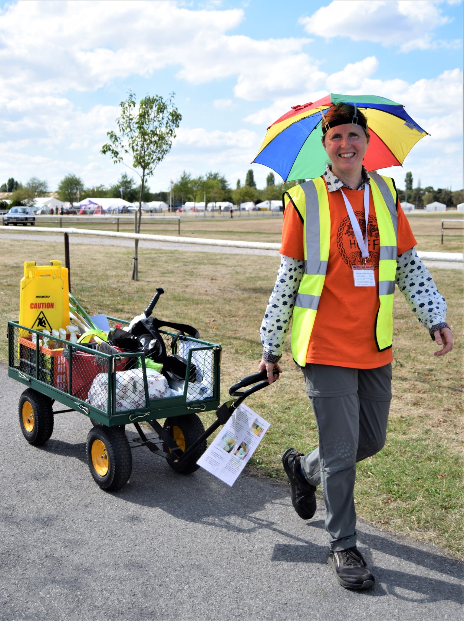 Photo of a woman pulley a trolley of cleaning supplies while wearing an orange HEFF T-shirt with a yelloe hi-vis waistcoat and an umbrella hat on her head