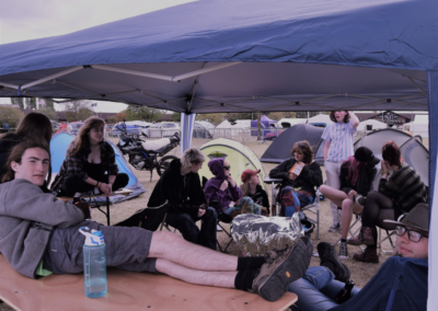 Photo of a group of teenagers sitting under a marquee shelter with a field of tents in the background