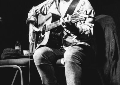 Black and white photo of a man sat on stage playing a guitar and singing into a microphone