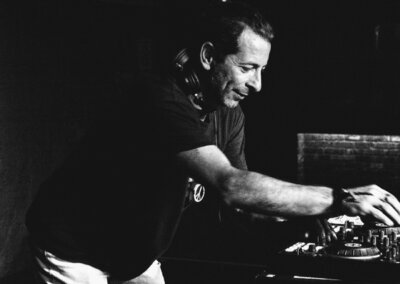 Black and white photo of a man wearing headphones round his neck while DJing at a turntable