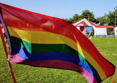 Close up photo of a rainbow pride flag being flown in a field with tents in the background