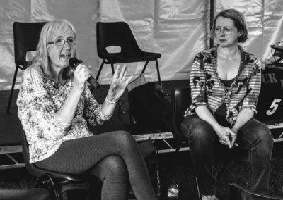 Black and white photo of two women sat on chairs in a marquee. One is speaking into a microphone