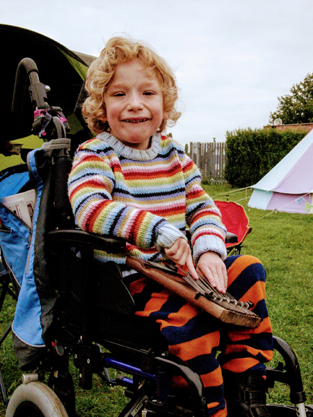 Photo of a young child wearing a bright stripy jumper and stripy trousers smiling for the camera while sitting in a wheelchair with tents in the background