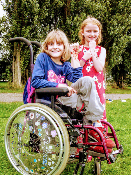 Photo of two young people with decorated arms and wrists. One of them is standing and the other is seated in a manual wheelchair decorated with stickers on the wheel. They are both smiling