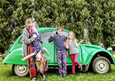 Photo of a family standing in front of a green Beatle car. The adult is holding one of the children upside down. The other two children are making peace signs with their hands