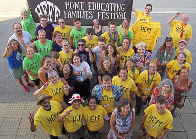 Group of people wearing yellow or green t-shirts featuring the HEFF logo. They are holding a large banner that reads "HEFF Home Educating Families' Festival".