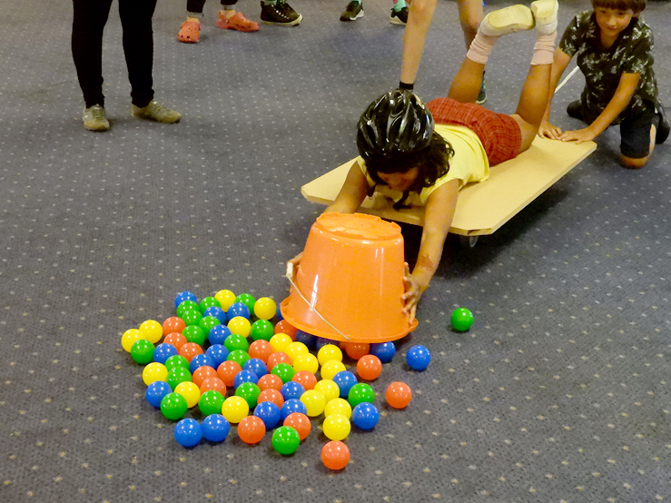 Photo of a young person wearing a black helmet while laying on a board on wheels and gathering balls into a large orange bucket