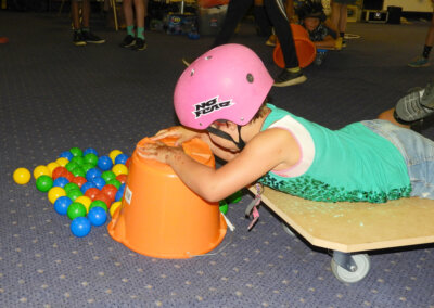 Photo of a young person wearing a pink helmet while laying on a board on wheels and gathering balls into a large orange bucket