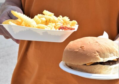 A close up of two paper plates being carried by a person in an orange tshirt. One has chips and the other has a cheeseburger.