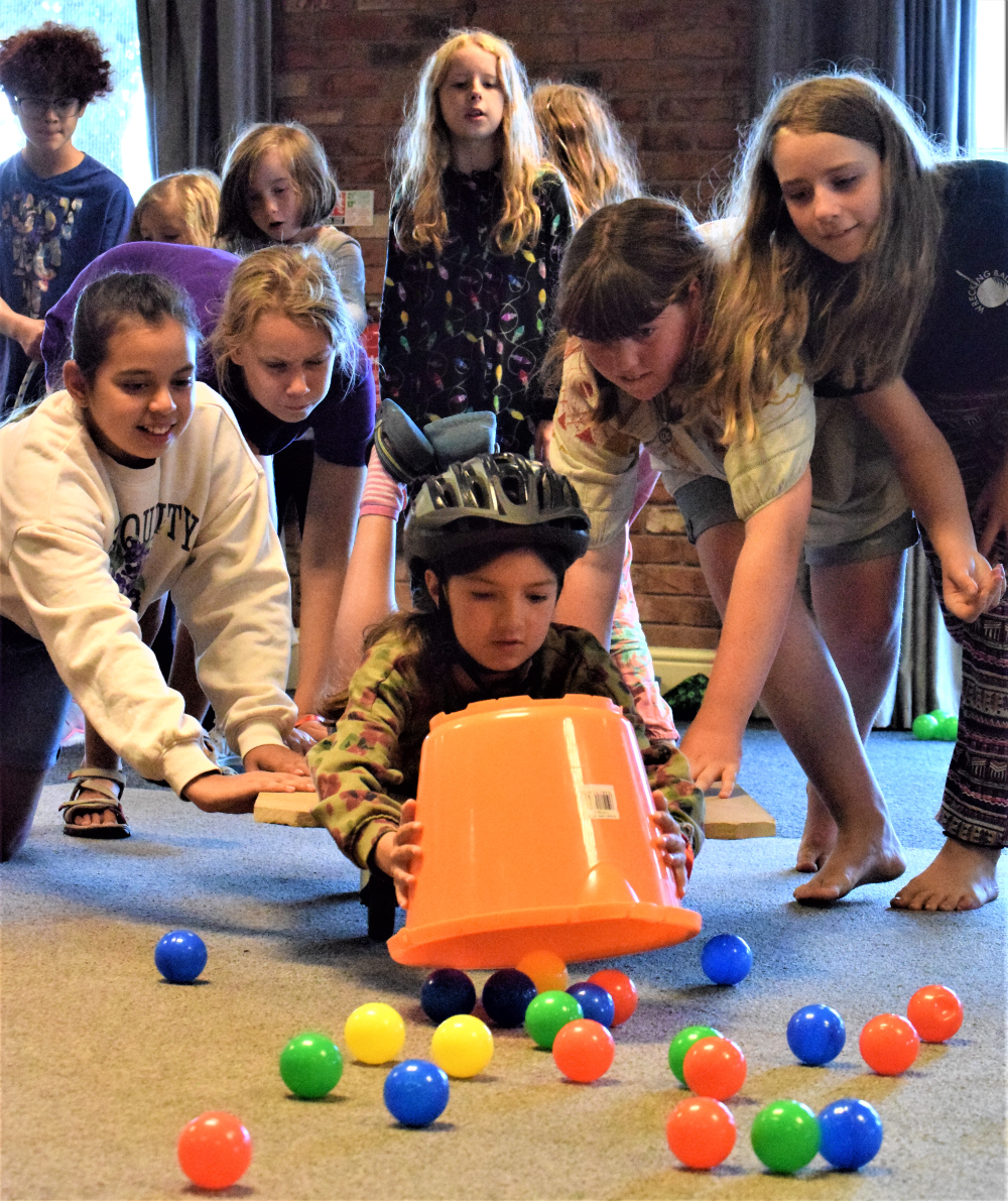 Photo of a young person wearing a black helmet while laying on a board on wheels and gathering balls into a large orange bucket with a group of young people behind them pushing the board