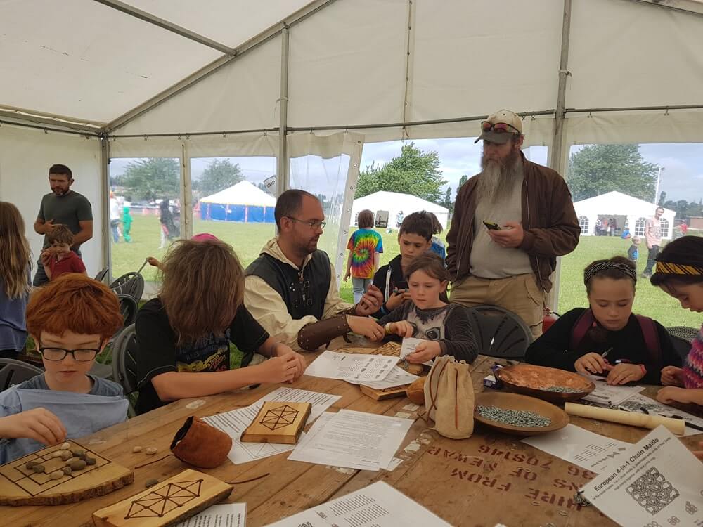 Photo of a group of people in a marque sitting at a table. The tabkle is covered in printed sheets of paper and pieces of wood with drawings on them